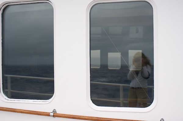 It’s difficult to capture the rolling swells and winds in a photograph, but I have been trying. We’ve had numerous rain squalls today and the winds have increased to 20 knots. The port side windows in this image created a reflection of my efforts and also a view to the starboard side of the ship.