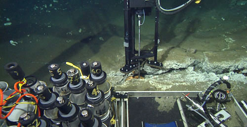 This view from the main camera on the remotely operated vehicle gives an idea of the various sampling tools carried to the seafloor to conduct chemistry studies. The laser Raman probe in a specially designed tripod with a hydraulic actuator sits above a fissure lined with white and orange bacteria. In the foreground, from left are the thermistor at the end of the orange cable, used to measure the temperature of venting fluids; push core tubes used to collect mud from the seafloor; and the pump and gauges used for the precise operation of the Raman spectrometer.