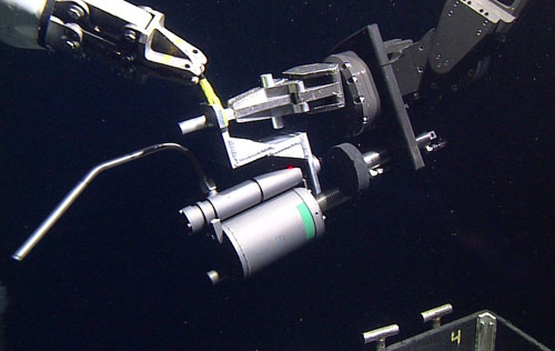 A sampler for collecting hot hydrothermal vent fluids is transferred from one manipulator arm's grip to the other prior to sampling. The nozzle on the left will be inserted into the black smoker's orifice. The trigger to draw in the sample is the hydraulic ram with the black circular plate attached below the manipulator's