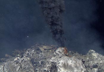 Hot, mineral rich water spews from a hydrothermal vent on the seafloor. Also known as black smoker, the mineral rich water appears as a column of black smoke rising from the seafloor. White, floculent bacterial mat is seen on the surface of the chimney below the black smoker.