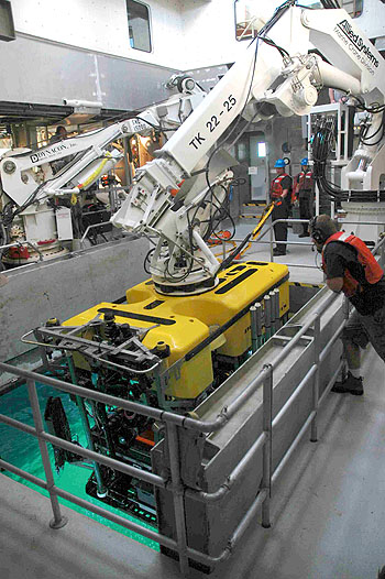 ROV Pilot Randy Prickett watches as the ROV Doc Ricketts is recovered from today's dive. There are two cranes in the moon pool: one for handling the tether management system during the dive (back crane operated by Dan Chamberlain) and another for launch and recovery of the vehicle (front crane operated by George Gunther). Jason Jordan and Matt Noyes are recovering the tether behind the vehicle.