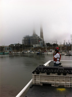 Second mate Miriam Anthony keeps watch on the back deck as the Western Flyer pulls out of from its slip in the harbor. The fog partly obscures the Moss Landing power plant’s stacks in the background.