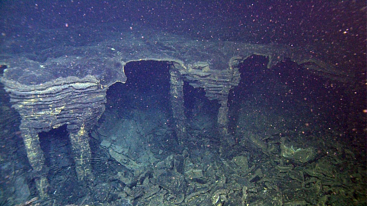 Narrow lava pillars miraculously support the thin roof of a drained lobate sheet flow near the fissure for this flow of the 2015 eruption. The lava channel for the flow (under the ROV) carried the lava down slope and out from under this roof, leaving the hollows we see and drainback veneers (the horizontal lines) on the lava pillars.