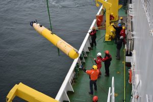Marine operations crew members recover a mapping robot from a mission in the Arctic Ocean. The crew members are standing on the green metal deck of a research ship on the right side of the frame. They are wearing hard hats, heavy work jackets, and dark pants. The robot on the left side of the frame has a thin cylindrical shape and is yellow with a black tail cone. The robot is connected to a nylon rope and crane off-screen at the top of the frame. The background is flat gray-blue ocean.