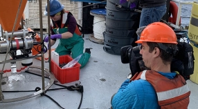 Woman collecting samples on ship deck with man in hard hat and life vest filming her.