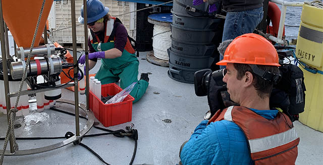 Woman collecting samples on ship deck with man in hard hat and life vest filming her.