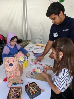 An MBARI intern (right) wearing a navy blue polo and white name tags helps two young visitors, right and left, seated at a table with a white tablecloth. The visitor on the left is wearing a purple-and-blue hooded sweatshirt and the visitor on the right is wearing a striped t-shirt. The visitors are located inside a white tent and are coloring white paper with crayons and assembling a sea otter puppet using a brown paper bag.