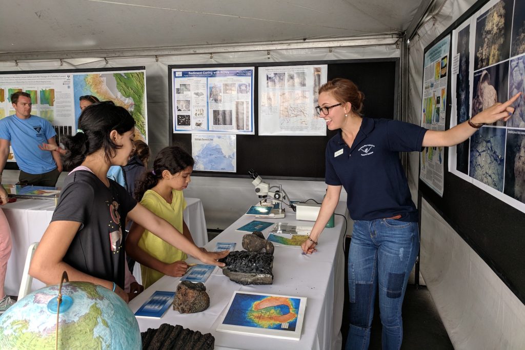 An MBARI staff member (right) wearing a navy blue polo shirt, jeans, and glasses is pointing to photos on a poster board while talking with two young visitors. One visitor is wearing a yellow shirt (center) and the second is wearing a gray shirt (left) and touching a rock sample on a table with a white tablecloth. In the background are other black poster boards with white posters and two Open House volunteers wearing blue t-shirts.