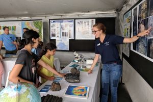 An MBARI staff member (right) wearing a navy blue polo shirt, jeans, and glasses is pointing to photos on a poster board while talking with two young visitors. One visitor is wearing a yellow shirt (center) and the second is wearing a gray shirt (left) and touching a rock sample on a table with a white tablecloth. In the background are other black poster boards with white posters and two Open House volunteers wearing blue t-shirts.