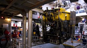 Members of MBARI’s marine operations prepare a robotic submersible for deployment. The submersible is in the center of the frame, with a yellow float and black metal instrumentation below. It is suspended from a white metal crane above the open silver metal doors of a moon pool. To the left is a crew member observing deployment, and to the right is a crew member operating the crane. In the background are various pieces of scientific and maritime equipment, illuminated by a skylight overhead.