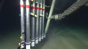 The black metal robotic arm of a deep-sea submersible places a silver metal cylinder covered in fine brown sediment into a rack of 12 sediment samples. The sediment samples are in tall, thin silver metal cylinders cradled by a short white plastic tube. The background is brown muddy seafloor fading to black water on the horizon.