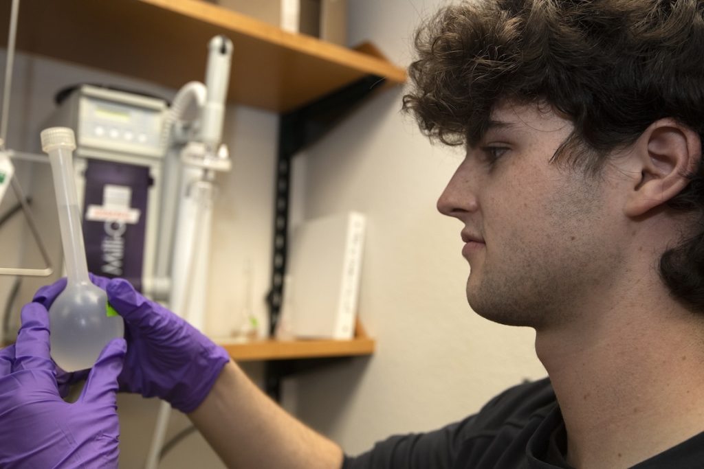 An intern holds up a clear plastic bottle to examine its contents. He is wearing purple gloves. In the background is a scientific instrument.