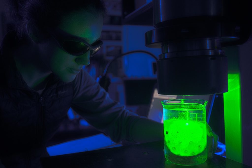 An intern wearing black goggles operates a laser imaging system in a dark laboratory. She is adjusting a glass beaker containing a deep-sea coral illuminated by bright green laser light.