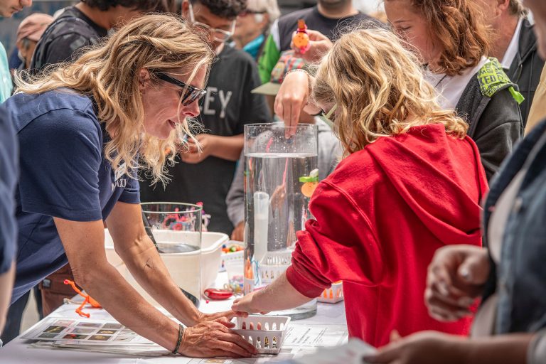 An MBARI researcher with shoulder-length blonde hair, black sunglasses, and a blue t-shirt holds a plastic box of supplies for a young visitor with shoulder-length blonde hair wearing a red hooded sweatshirt. The two are outdoors on a sunny day with other visitors and a science demonstration in the background.