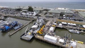 A drone flies above MBARI’s Open House. Crowds of visitors walk around several large white tents set up in the parking lots and dock adjacent to MBARI’s research facilities. In the background are gray concrete buildings with green roofs, brown sandy beach, grayish-blue ocean, and blue sky. In the foreground is Moss Landing Harbor with greenish-brown water, a gray metal research ship, a blue boat, and a white boat.