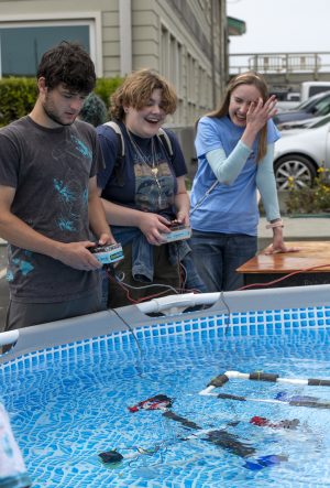 Two visitors at the MBARI Open House pilot a pair of small underwater robots in a blue pool of water. The visitor on the left has short black hair and is wearing a gray t-shirt and gray pants. The visitor on the right has shaggy brown hair and is wearing a tan backpack, a blue graphic t-shirt, and olive-green pants with a denim jacket tied around their waist. On the right is an Open House volunteer with long blonde hair wearing a light blue t-shirt and jeans. In the foreground is a portable swimming pool with a blue-and-white checkered lining filled with water and two underwater robots made of white plastic pipe, black foam, red propellers, and blue propellers. In the background is a tan building and several parked cars.