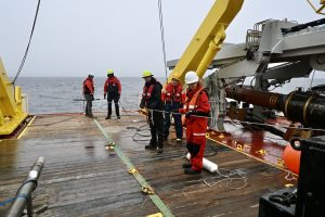 Five researchers prepare to deploy a scientific instrument from the stern of a research ship on an overcast day. The researchers are wearing hard hats, orange life vests, orange jackets, black pants, and dark boots. They are holding a line connected to scientific instruments out of frame to the right. They are standing on the wooden deck of the ship with maritime equipment on the right side of the frame. The background is gray ocean and overcast gray sky.