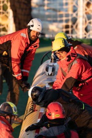 Six marine operations crew prepare to deploy a seafloor mapping robot from the deck of a research ship on a sunny day. The crew are all wearing hard hats and bright orange jackets. The robot has a cylindrical yellow plastic housing. In the background is the green deck of the ship with assorted maritime equipment.