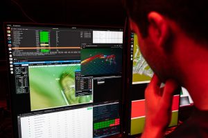 An engineer examines a live feed of navigation data from sensors aboard a deep-sea robot. The engineer has short black hair and is wearing a blue t-shirt. He is watching a computer screen with several windows displaying technical data, computer code, and navigation plots. The frame is illuminated with red light.