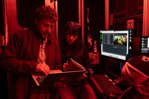 Two scientists review scientific data on a laptop computer screen. The researcher on the left is standing, has short curly brown hair, is wearing a dark fleece jacket and a plaid shirt, and is holding a silver metal laptop computer. The researcher on the right is seated, has long brown hair, is wearing a dark jacket and jeans, and is holding a notebook and pen. The researchers are in the control room aboard a research ship with computer monitors displaying a live camera feed and data from a deep-sea robot. The frame is illuminated with red light.