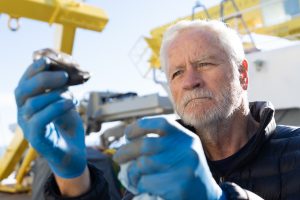 A scientist measures a piece of ice on the deck of a research ship on a sunny day. The scientist has white hair and a white beard and is wearing a blue down jacket and bright blue rubber gloves. He is holding a pair of silver metal calipers. In the background is yellow metal machinery aboard the ship and clear blue sky.