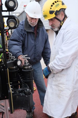 Two scientists examine sediment samples collected by a deep-sea robot. The scientist on the left has a white beard and is wearing a white hard hat, a blue jacket, blue jeans, and brown shoes. The scientist on the right has a gray goatee and is wearing a yellow hard hat, white lab coat, and bright blue rubber gloves. They are examining black metal canisters containing cylindrical push cores with silver metal handles. In the background is the deck of the research ship.