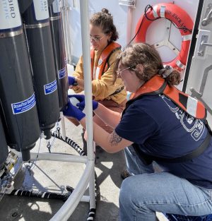 Two researchers collect water samples from a scientific instrument on the deck of a research ship on a sunny day. The researcher in the foreground has long brown hair in a bun and is wearing glasses, purple gloves, a navy-blue graphic t-shirt, an orange life vest, and blue jeans. The researcher in the background has brown hair in a bun and is wearing glasses, purple gloves, a yellow sweatshirt, an orange life vest, and red pants. Both researchers are sitting on blue plastic crates. To the left side of the frame is a scientific instrument with a white metal frame and a carousel of gray plastic bottles. In the background is the white metal wall of a research ship with an orange life ring.