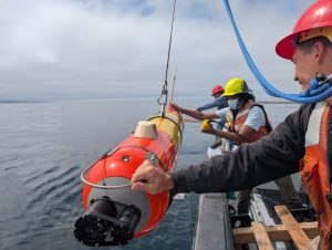 Three engineers deploy an underwater robot. The engineers are wearing hard hats and orange life vests and standing on the deck of a small boat on the right side of the frame. The robot on the left side of the frame has an orange-and-yellow housing and is outfitted with two black cameras. The robot is suspended from a crane out of frame. In the background are flat grayish-blue ocean and gray cloudy sky.