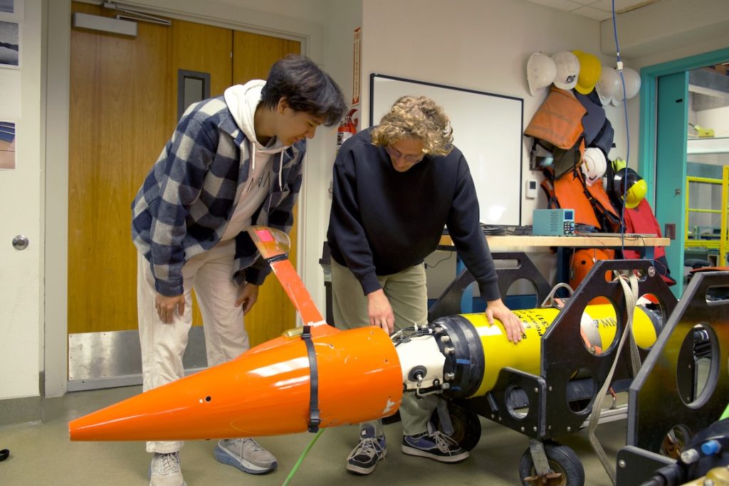 Two interns disassemble an autonomous robot in a brightly lit engineering lab. The robot has a yellow plastic housing with an orange plastic tail cone, and is sitting on a black plastic cart.