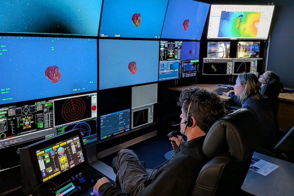 A submersible pilot and two biologists sit in black leather chairs and watch video monitors in the dark submersible control room aboard a research ship. The pilot is in the foreground. He has wavy salt-and-pepper hair, is wearing a headset, a black hooded sweatshirt, and gray pants, and is holding a black rubber joystick. The biologist in the center has short blonde hair. She is wearing a headset and a navy-blue jacket. The biologist in the background has short blonde hair. She is wearing glasses and a headset. All three are watching a wall of video monitors on the left side of the frame displaying a video feed and data from the deep sea. A bright red comb jelly swimming in open blue water is displayed on four of the monitors at the center of the wall. A colorful topographic map is displayed on a monitor on the right.
