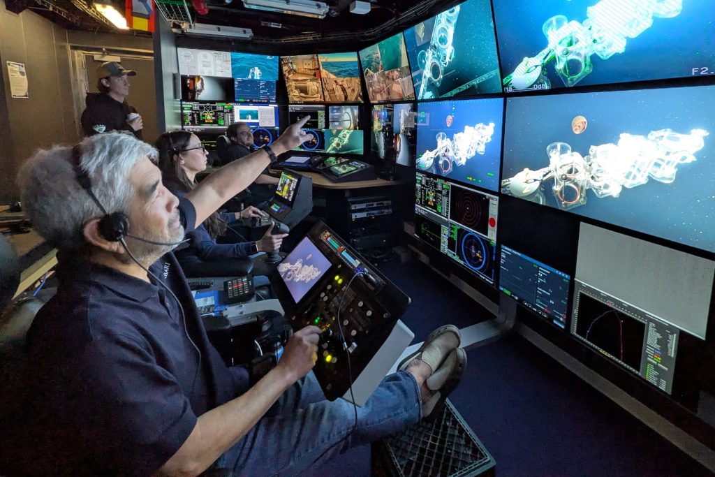 An MBARI researcher points to a wall of video monitors in the dark submersible control room of a research ship. The researcher is seated in a black leather chair with his legs up on a black plastic milk crate. In the background are two submersible pilots seated in chairs and operating submersible controls, and a third submersible pilot holding a cup of coffee. The video monitors on the right side of the frame display a live feed from a deep-sea submersible, showing an array of clear plastic sampling canisters and a bright red jelly.