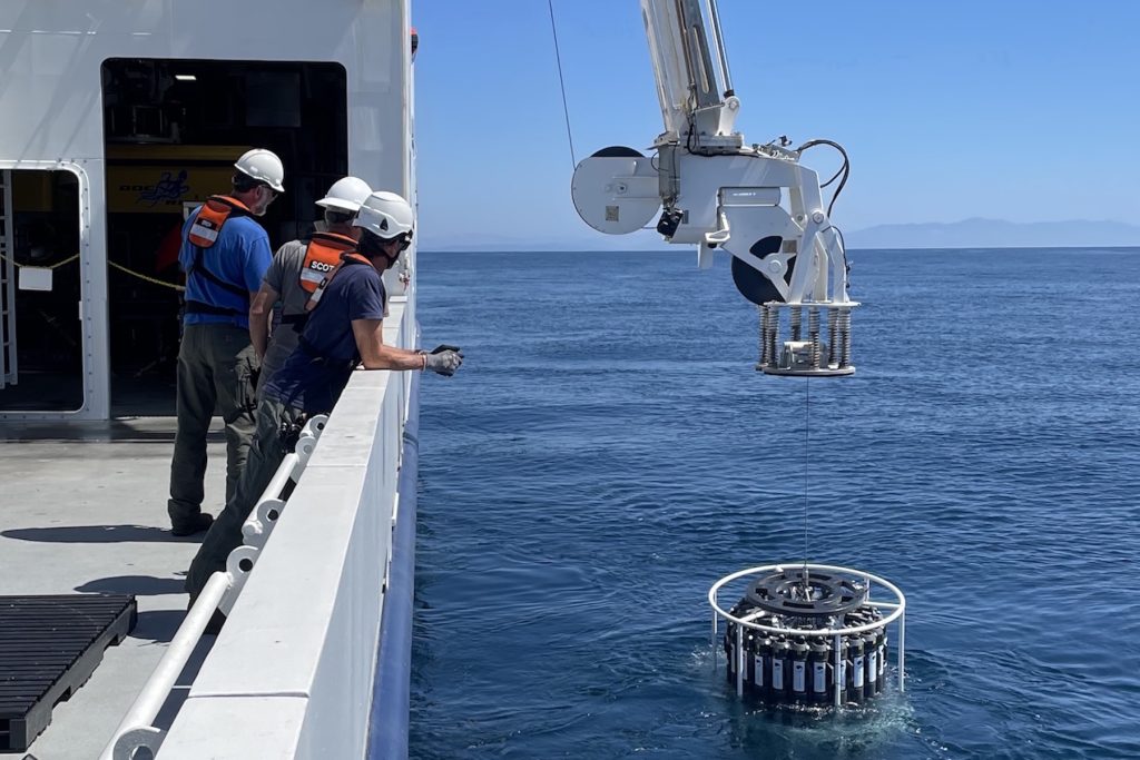 Marine operations crew deploy a scientific instrument over the side of a research ship. On the left side of the frame, three crew members wearing white hard hats, orange life vests, blue shirts, and dark pants stand on the gray metal deck of a ship, leaning against a white metal railing. On the right side of the frame, a white metal crane lowers a round scientific instrument with a carousel of gray plastic bottles. In the background are still blue ocean and blue sky, with hazy clouds and coastal mountains on the horizon.