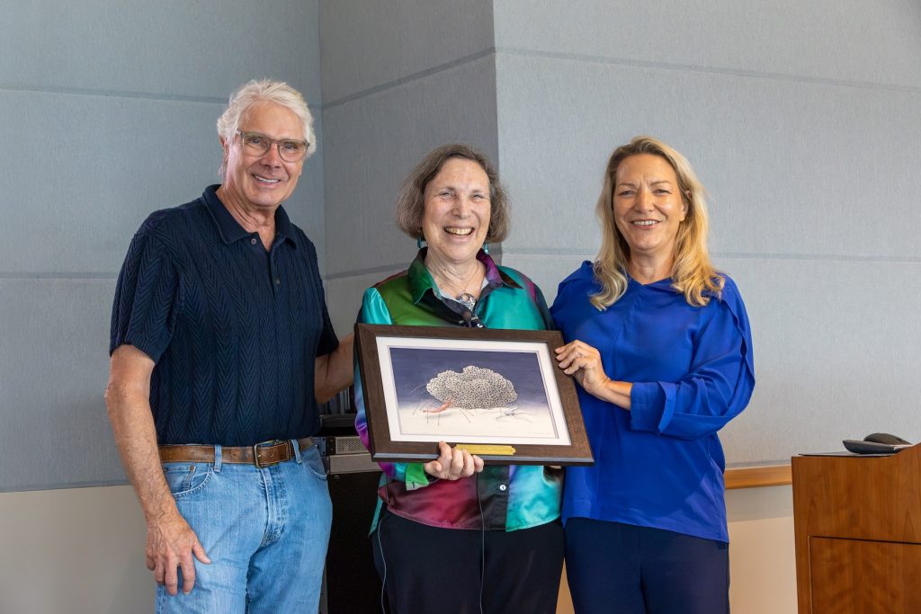 Three scientists pose for a photo on a raised stage in a large conference room. The scientist in the middle is holding a framed print of an illustration of a deep-sea organism.