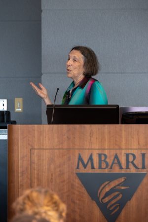 A scientist standing behind a wooden podium gives a presentation to an audience seated in a large conference room.