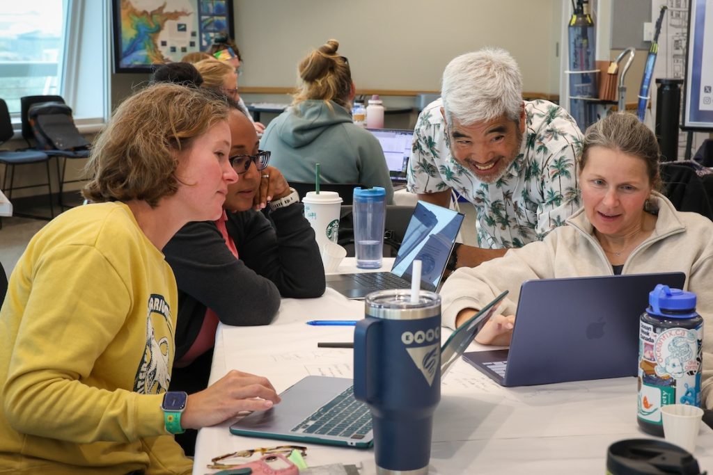 A scientist speaks with three teachers during an education workshop. He is crouched down while a teacher shows him something on a computer screen. In the background is a crowded room full of teachers seated around tables for an interactive small-group activity.