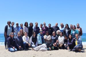 Thirty workshop participants and staff pose for a photo on a sunny day at the beach. In the background is greenish-blue ocean with clear blue sky on the horizon.