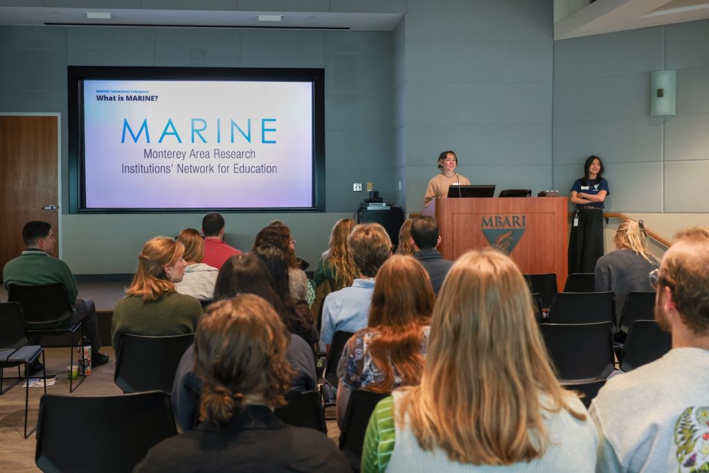 Two presenters stand behind a podium in a large seminar room welcoming attendees to a networking and professional development workshop.