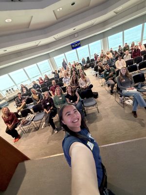 A selfie photo shows an organizer and a large audience of participants gathered for a workshop in a large seminar room.