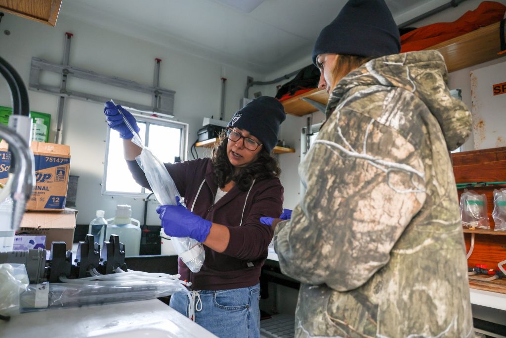 Two summer interns process water samples in the lab aboard a research ship. The intern on the left is wearing purple gloves while transferring water from a thin glass tube into a large plastic tube inside a clear plastic bag. In the background are shelves of scientific equipment.