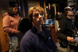 A summer intern wearing a black headset watches a video feed from a robotic submersible from the submersible control room aboard a research vessel. He is seated in a tall black chair, with a submersible pilot seated next to him operating a joystick. In the background is an engineer standing in front of electronic equipment.
