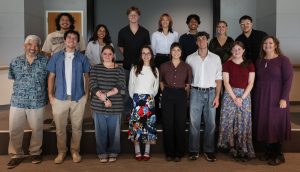 A group of 13 summer interns and two program staff pose for a photo in a conference room. The group is arranged into two rows, with one row standing on a raised stage in front of a projection screen.
