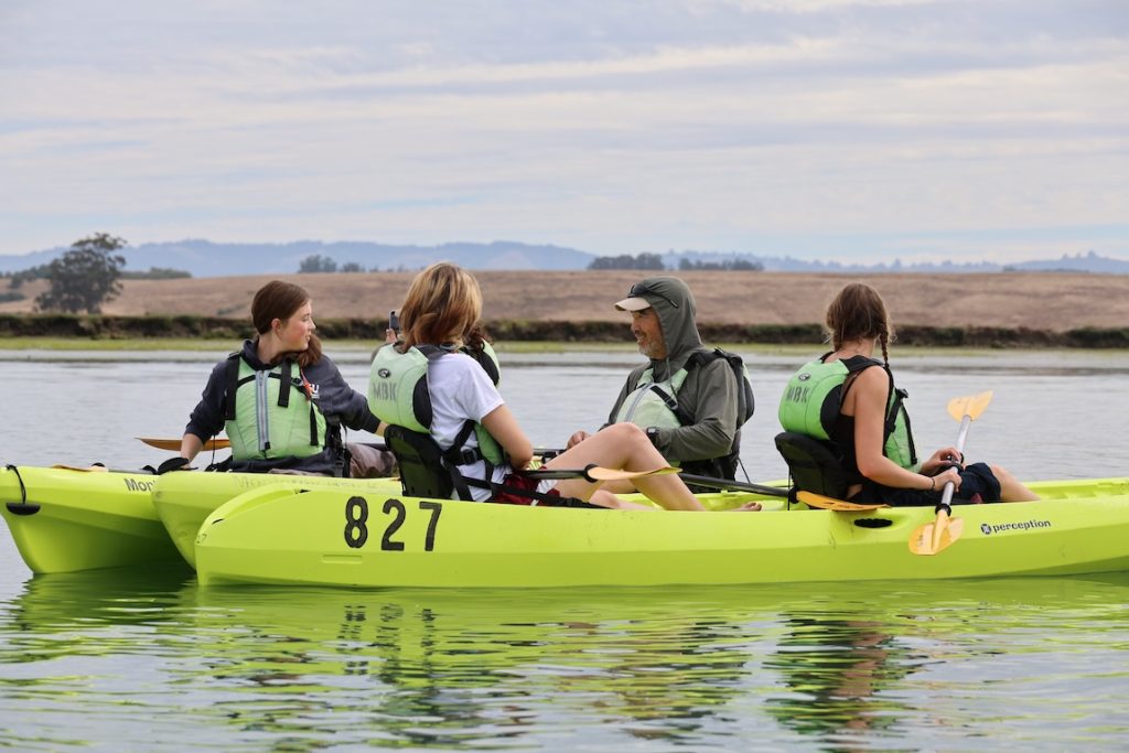 A scientist talks with four summer interns during a kayaking excursion. He has parked his bright yellow kayak between two other kayaks in still water in coastal wetlands. In the background is the riverbank with brown hills and green trees, with coastal mountains and hazy overcast sky on the horizon.