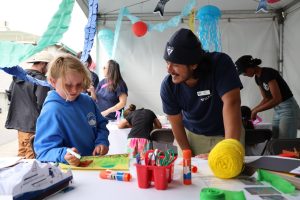 A summer intern and a young child standing outdoors under a large white tent are making ocean-inspired crafts at the MBARI Open House.