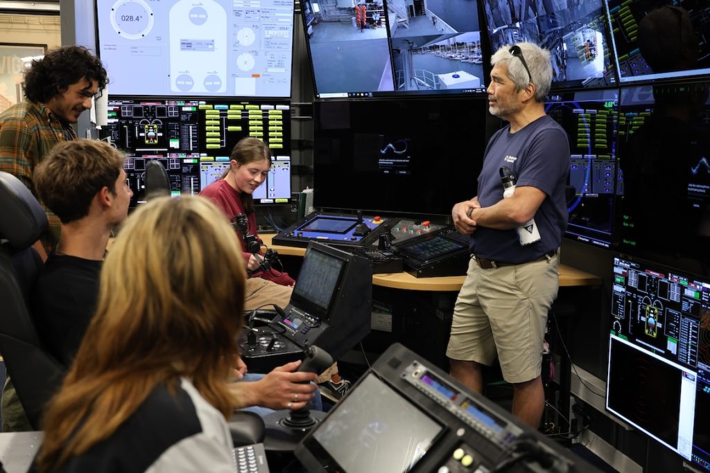 A scientist speaks with four summer interns in the submersible control room aboard a research ship. He is standing in front of monitors displaying live video feeds and data from the ship’s internal cameras. The interns are seated in chairs on the left side of the frame.