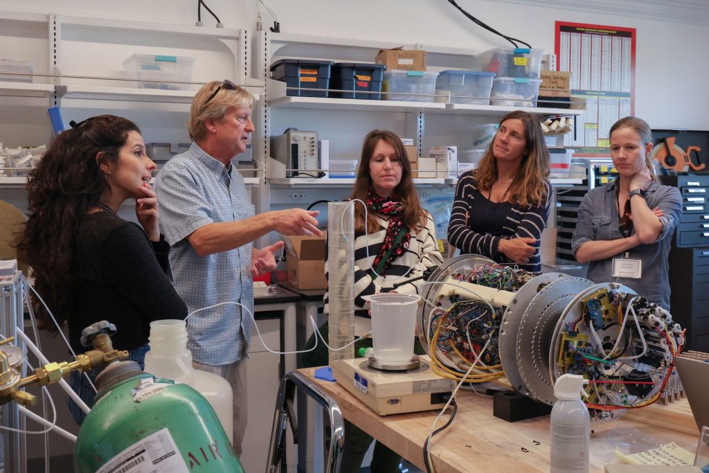 A scientist tours a group of workshop participants through a research laboratory. The scientist has short blond hair and is wearing sunglasses, a blue short-sleeved shirt, and khaki pants. There is one workshop participant on his right and three participants on his left. He is pointing to a cylindrical silver metal scientific instrument covered in wires and electronics that is assembled on a wooden countertop. In the background are white walls and shelves of containers full of instrument components.