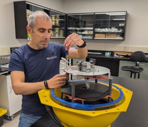 A scientist prepares a scientific instrument in a research lab. The scientist is screwing components onto a silver metal plate on a gray plastic platform inside an open yellow plastic housing. In the background are the black counters, black cabinets, and white walls of a research lab.