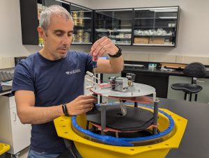 A scientist prepares a scientific instrument in a research lab. The scientist is screwing in components on a silver metal plate on a gray plastic platform inside an open yellow plastic housing. In the background are the black counters, black cabinets, and white walls of a research lab.