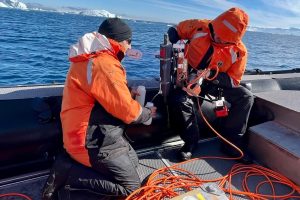 Two researchers wearing orange jackets and black nylon pants process water samples aboard a black Zodiac boat. The researcher on the right is holding a gray canister of water and a spool of orange cable. The researcher on the left is collecting water from the gray canister in two white plastic bottles. In the background are blue water and blue sky with large white icebergs on the horizon.