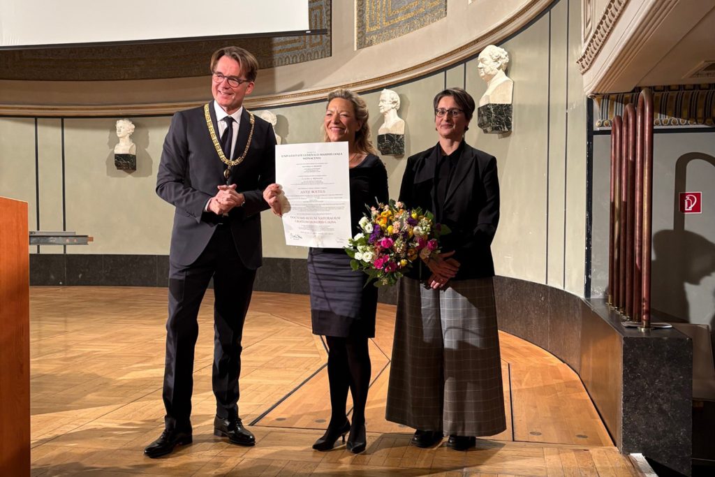 Three people stand in a room with wooden floors and a curved wall behind them. A smiling man with brown hair stands on the left. He is wearing a suit, a tie, white shirt. The blonde woman in the middle is holding a large white rectangular card. She is wearing a dark knee-length skirt, dark tights, and shoes. The woman on the right has brown hair and is wearing a black jacket and a dark gray long skirt. She is holding a bouquet of flowers.