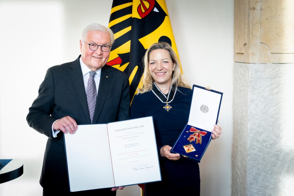 German Federal President and MBARI President and CEO pose at a government medal ceremony. The German Federal President is standing on the left holding an official proclamation letter. MBARI’s President and CEO is on the right, holding her medal in a blue box. In the background are white walls and a flag.